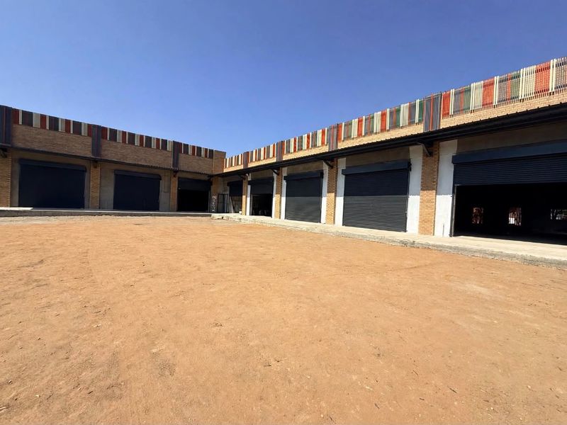 Exterior view of a commercial retail property featuring multiple roller shutter doors, brick facade detailing, and a large unpaved yard area.
