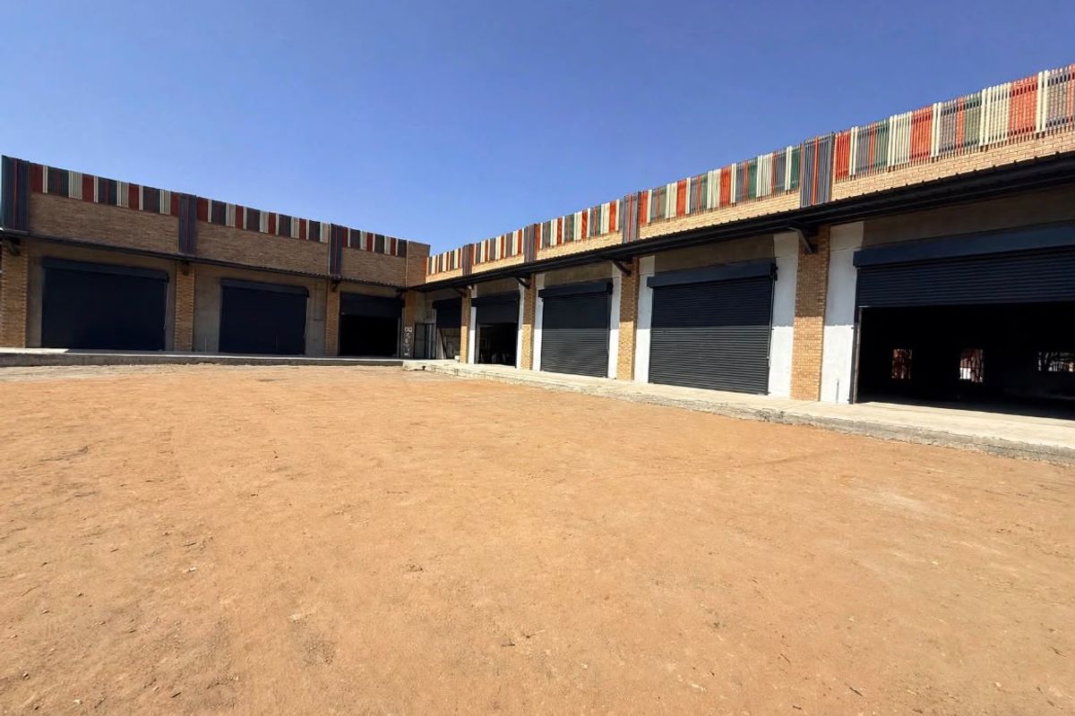 Exterior view of a commercial retail property featuring multiple roller shutter doors, brick facade detailing, and a large unpaved yard area.