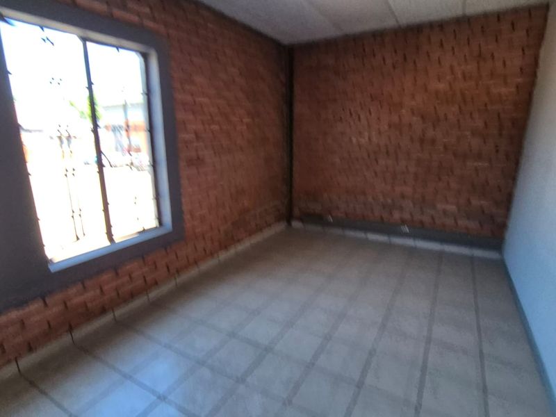 An empty office space featuring exposed brick walls, tiled flooring, and windows with security bars, offering a functional industrial aesthetic.