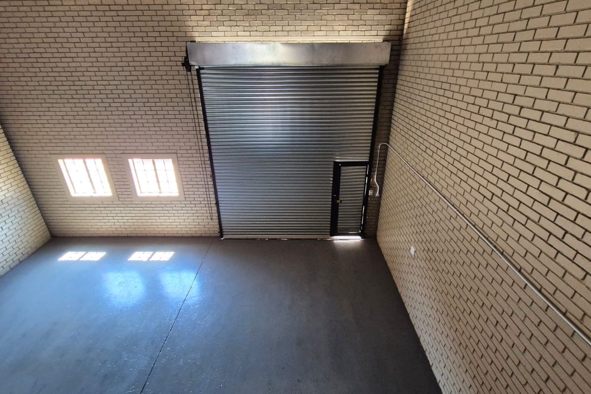 Spacious industrial utility area featuring high-clearance roller shutter door, durable concrete flooring, and exposed brick walls, suitable for storage or workshop use.