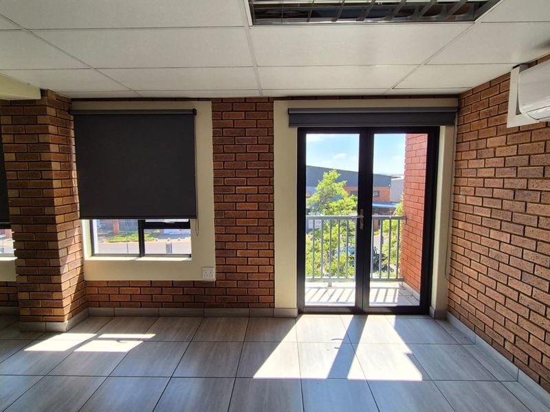 A bright, empty office space featuring exposed brick walls, tiled flooring, a drop ceiling, and direct access to a small balcony.