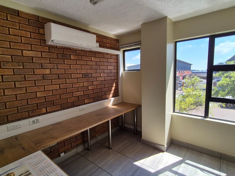A bright, functional office space featuring an exposed brick feature wall, built-in desk, split-system air conditioning, and large windows providing natural light.