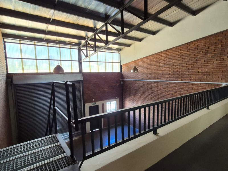 Spacious industrial utility area featuring exposed brick walls, high metal roof trusses, a roller shutter door, and a mezzanine level with metal railing.