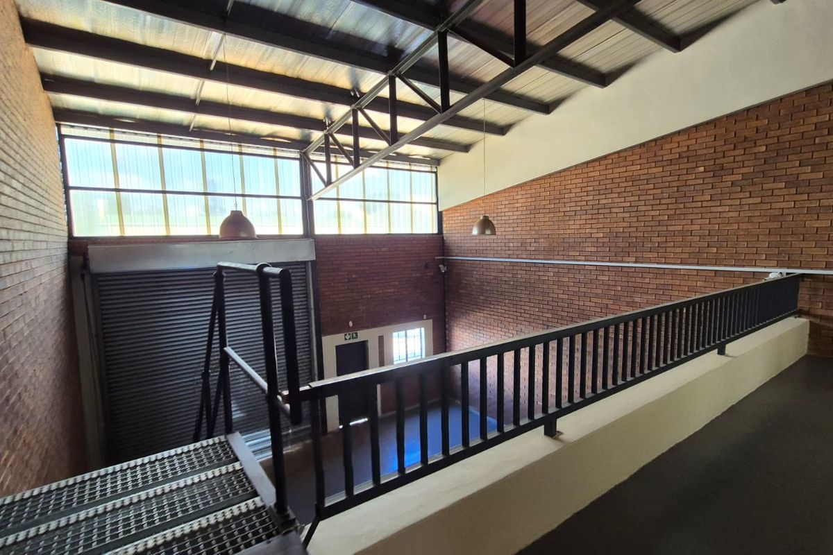 Spacious industrial utility area featuring exposed brick walls, high metal roof trusses, a roller shutter door, and a mezzanine level with metal railing.
