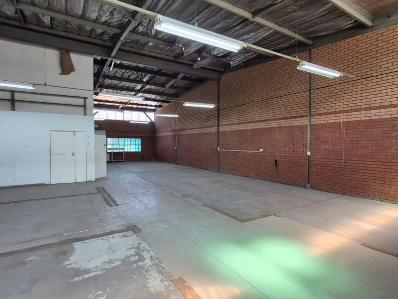 Spacious industrial warehouse unit featuring exposed brick walls, concrete flooring, high ceilings with steel trusses, and functional fluorescent lighting throughout.