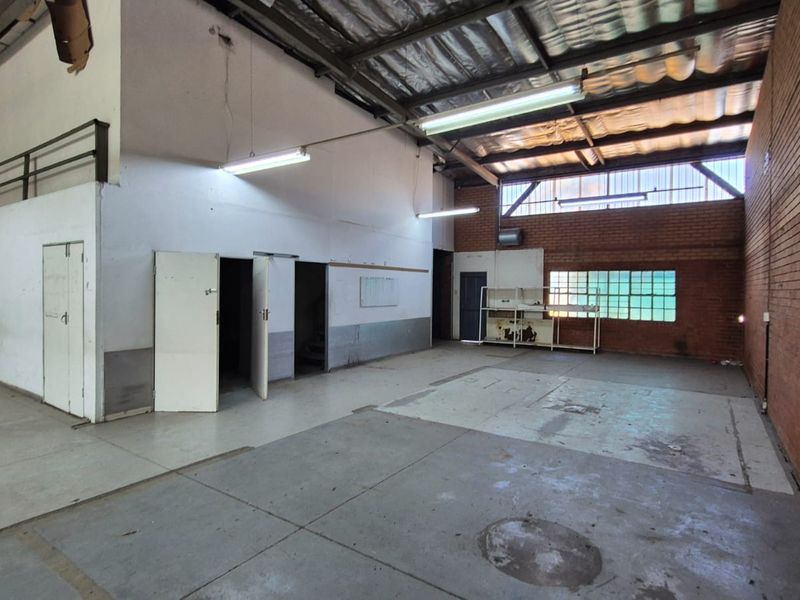 Spacious industrial utility area featuring exposed brick walls, high steel-truss ceilings, fluorescent lighting, and durable concrete flooring, suitable for workshop or storage use.