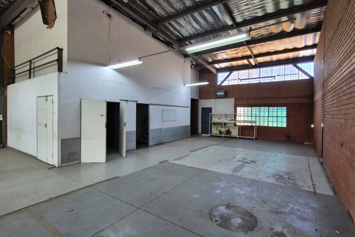 Spacious industrial utility area featuring exposed brick walls, high steel-truss ceilings, fluorescent lighting, and durable concrete flooring, suitable for workshop or storage use.