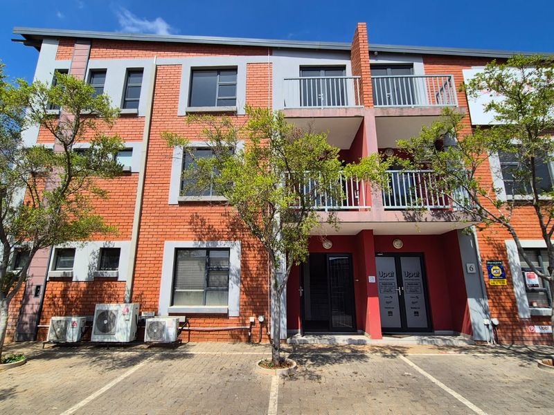Front elevation of a multi-story industrial or commercial building featuring red brick facade, balconies with metal railings, and dedicated paved parking.