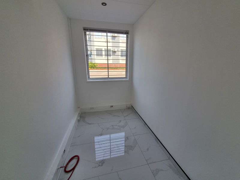A compact, empty office space featuring modern marble-look floor tiles, a window with security bars, and recessed lighting.