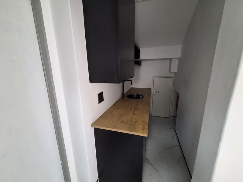 A compact, modern scullery or kitchenette area featuring dark cabinetry, a wooden countertop, integrated sink, and light-colored tiled flooring.