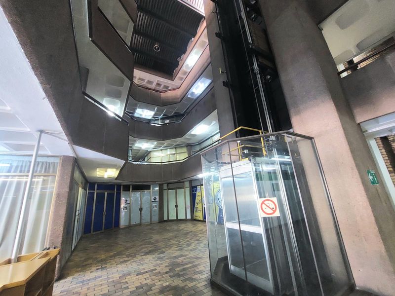A multi-level office atrium featuring a central glass elevator, brick flooring, and open-plan corridors overlooking the main lobby area.