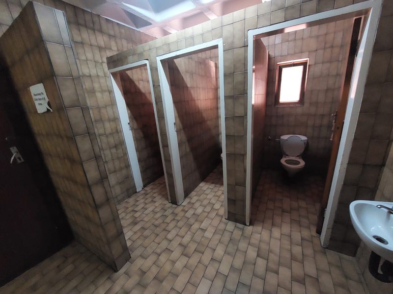 A communal office restroom featuring multiple toilet cubicles with dated ceramic wall and floor tiling and a wall-mounted basin.