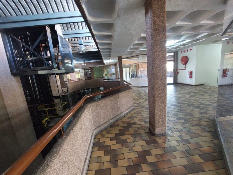 Spacious office building atrium featuring a central elevator shaft, textured concrete columns, and tiled flooring with a unique waffle-patterned ceiling design.