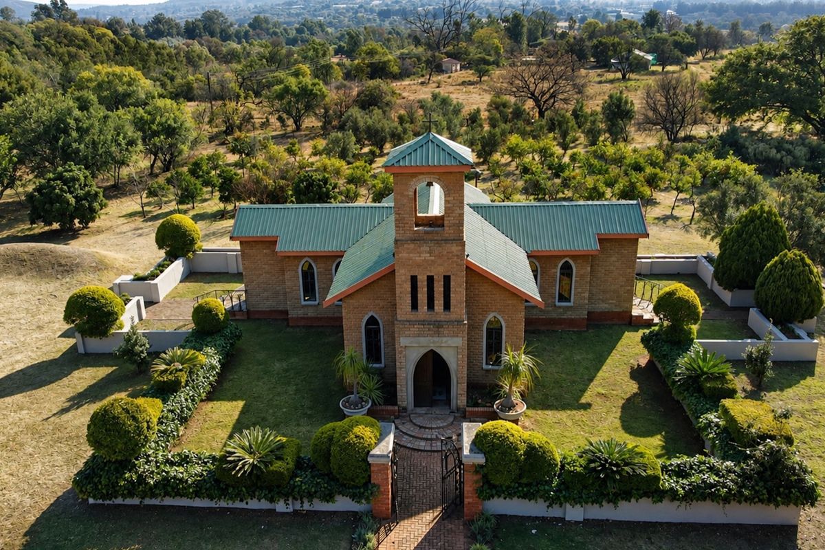 Aerial view of a charming brick chapel featuring a metal roof, bell tower, and arched windows, set within a well-maintained landscaped garden.