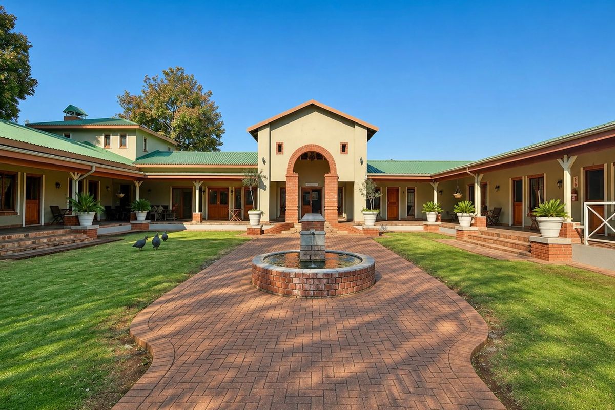 A well-maintained courtyard featuring a central brick fountain, surrounded by a covered veranda and a U-shaped building structure with green metal roofing.