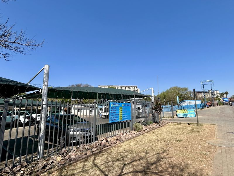 Street-level view of a commercial property featuring shaded parking bays, perimeter steel fencing with razor wire, and paved sidewalk access.