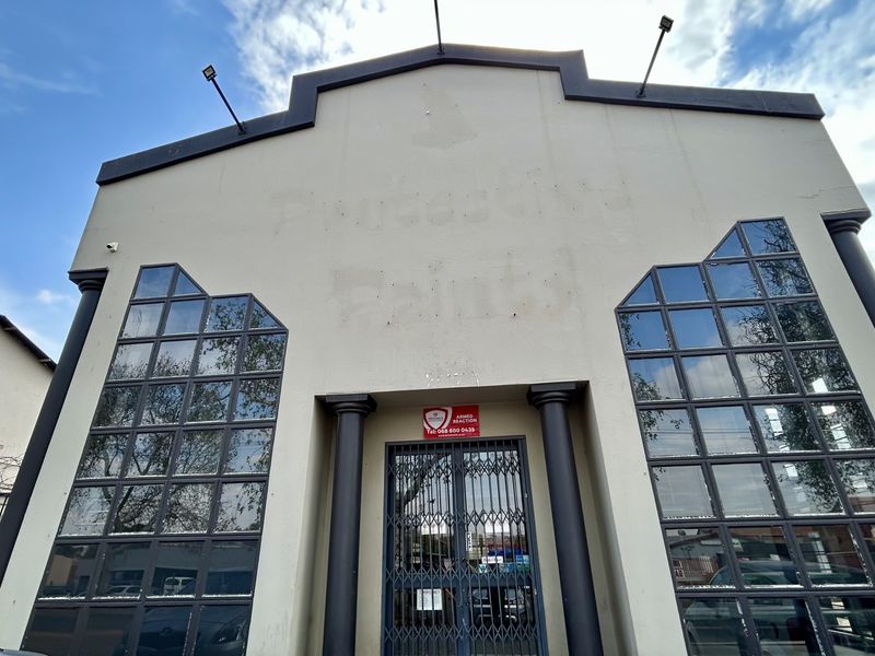Front facade of an industrial building featuring decorative columns, large multi-pane windows, and security infrastructure including a metal gate and floodlights.