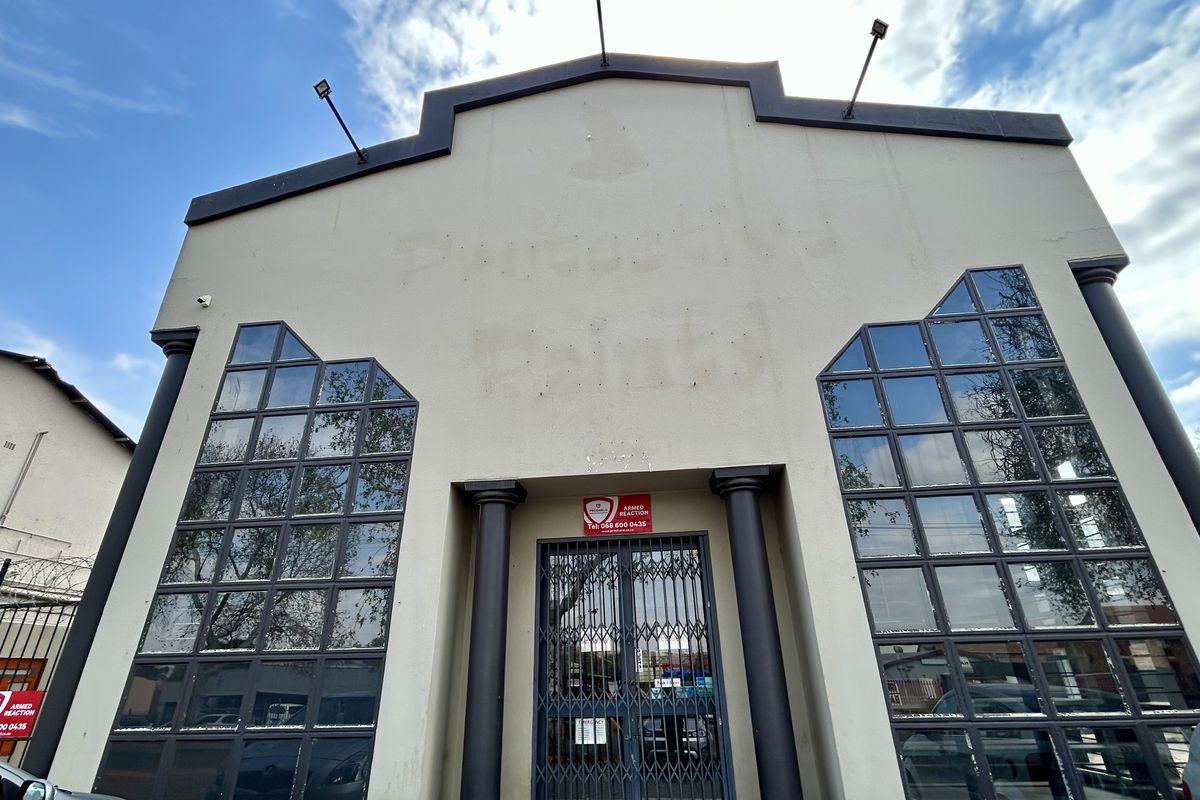 Front facade of an industrial building featuring decorative columns, large multi-pane windows, and security infrastructure including a metal gate and floodlights.