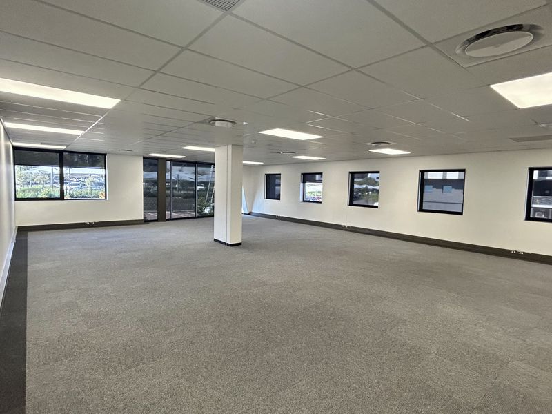 A spacious, open-plan office area featuring neutral carpet tiles, a suspended ceiling with recessed lighting, and large windows providing ample natural light.