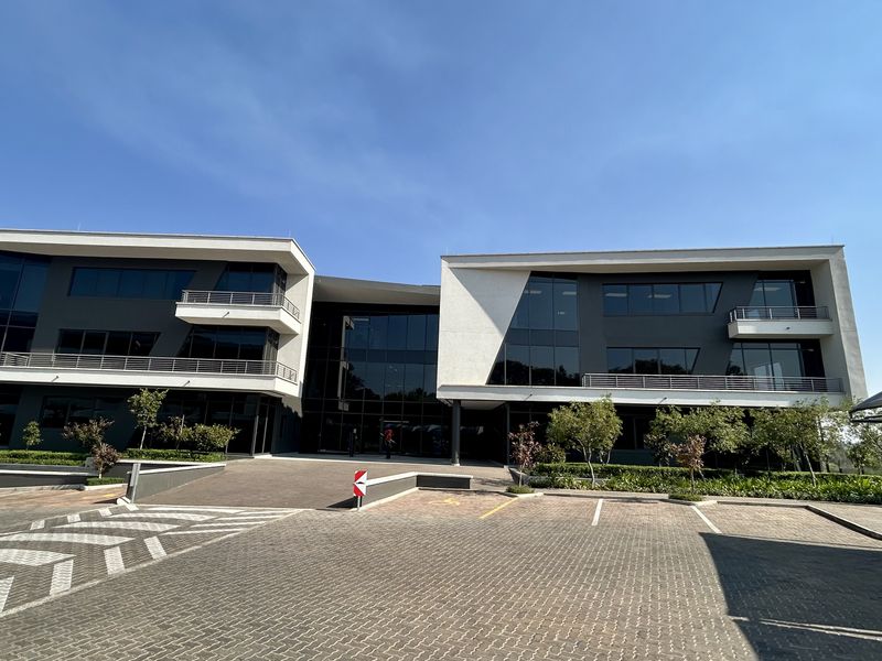Modern multi-story office building featuring a glass facade, integrated balconies, and a paved parking area with landscaped surroundings.