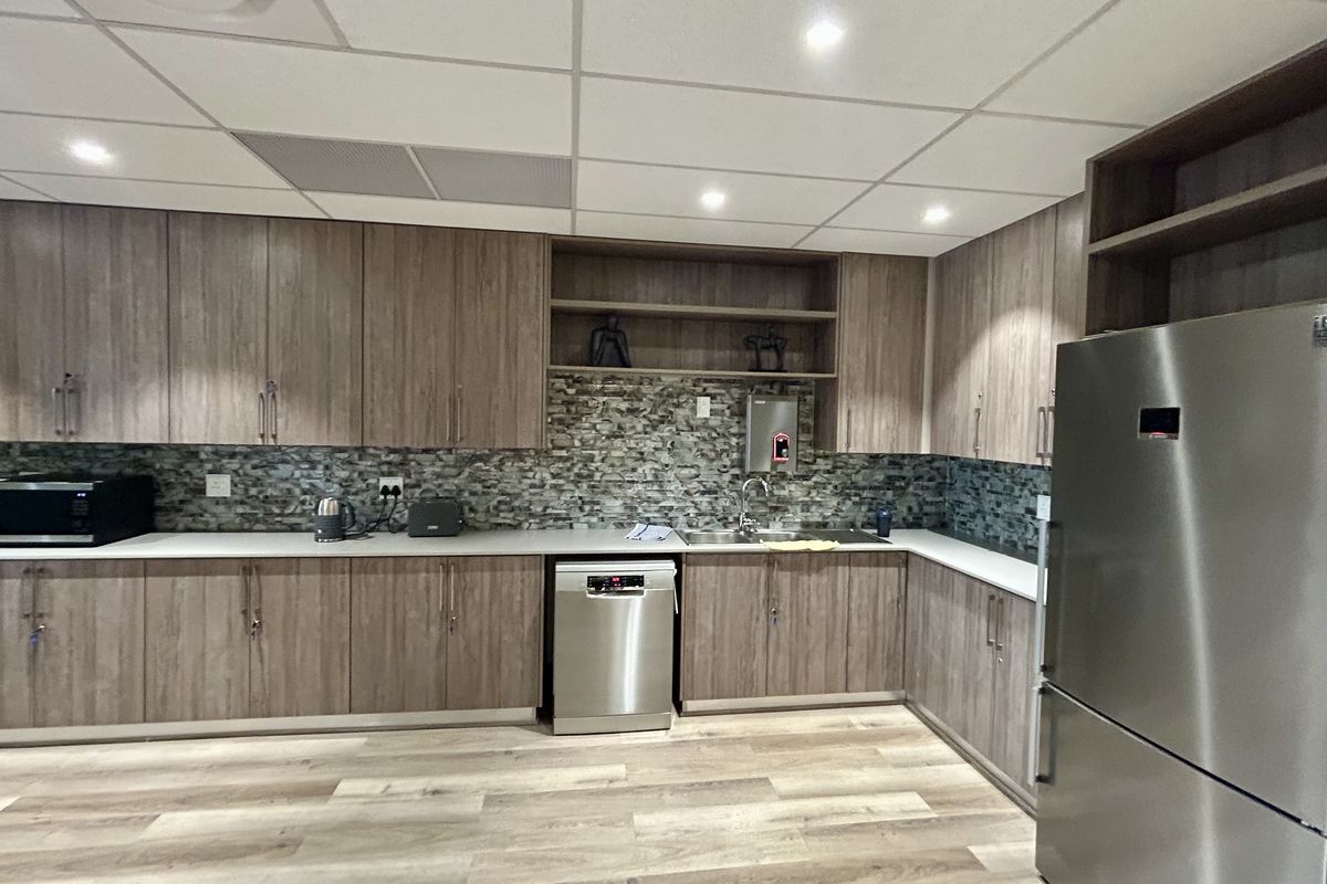 A modern office kitchen featuring wood-grain cabinetry, a stone-look backsplash, integrated dishwasher, and durable laminate flooring with recessed ceiling lighting.