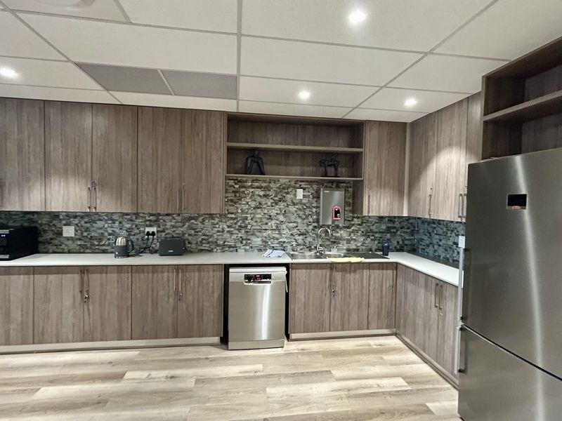 A modern office kitchen featuring wood-grain cabinetry, a stone-look backsplash, integrated dishwasher, and durable laminate flooring with recessed ceiling lighting.