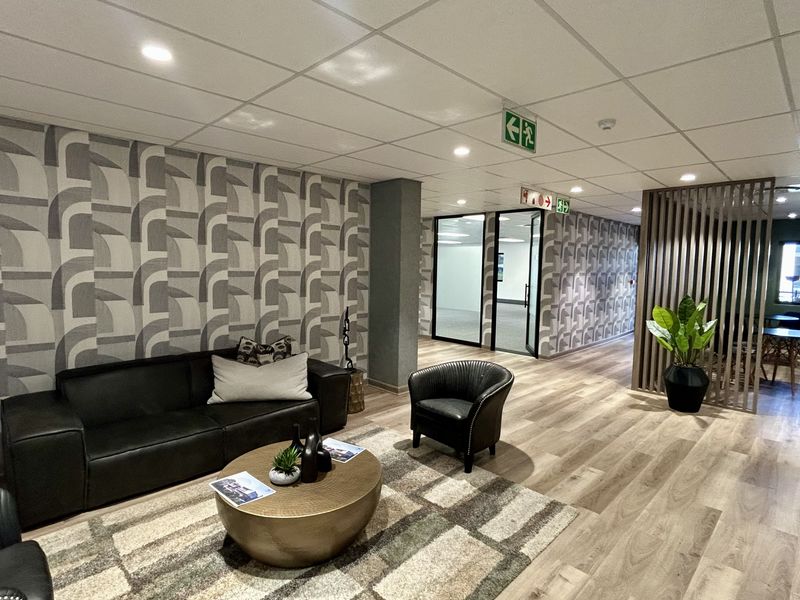 Modern office reception or breakout area featuring wood-look flooring, patterned feature wallpaper, suspended ceiling with recessed lighting, and a contemporary timber slat room divider.