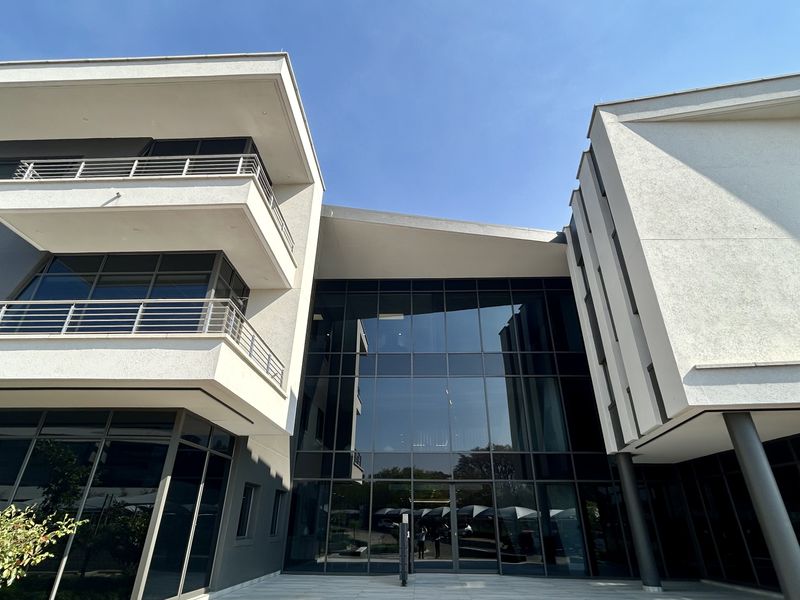 Modern office building exterior featuring a prominent glass curtain wall entrance, multi-level balconies with metal railings, and a clean, contemporary architectural design.