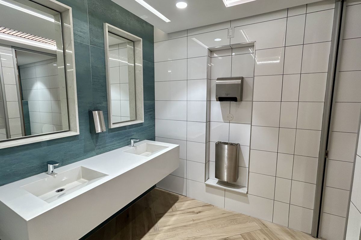 Modern office bathroom featuring a sleek double vanity, contemporary tiled walls, recessed lighting, and integrated wall-mounted hand dryer and waste disposal unit.
