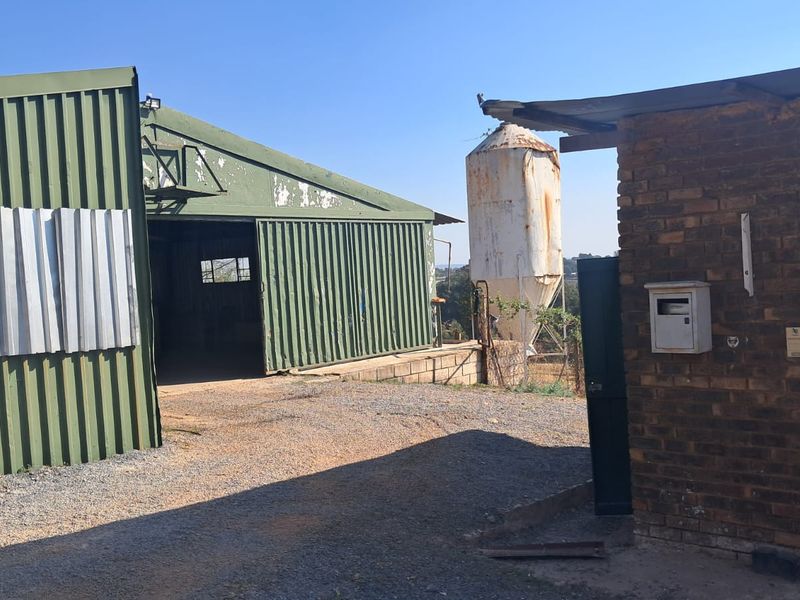 Industrial yard area featuring a large corrugated metal warehouse structure, an external silo, and a brick utility building with gravel surfacing.