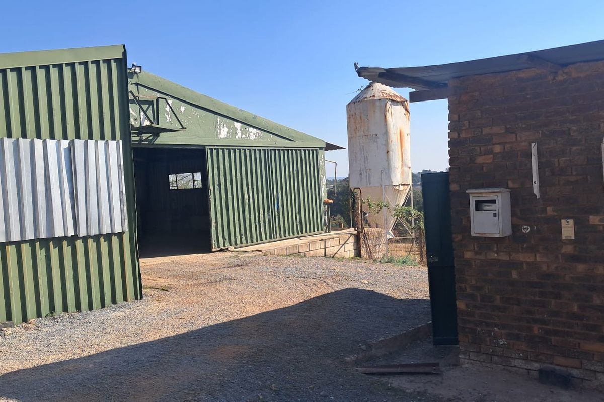 Industrial yard area featuring a large corrugated metal warehouse structure, an external silo, and a brick utility building with gravel surfacing.