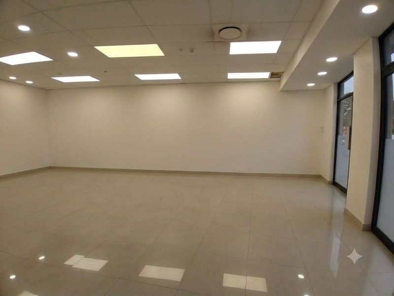 A spacious, empty open-plan retail unit featuring polished tiled flooring, a suspended ceiling with recessed lighting, and a glass entrance door.
