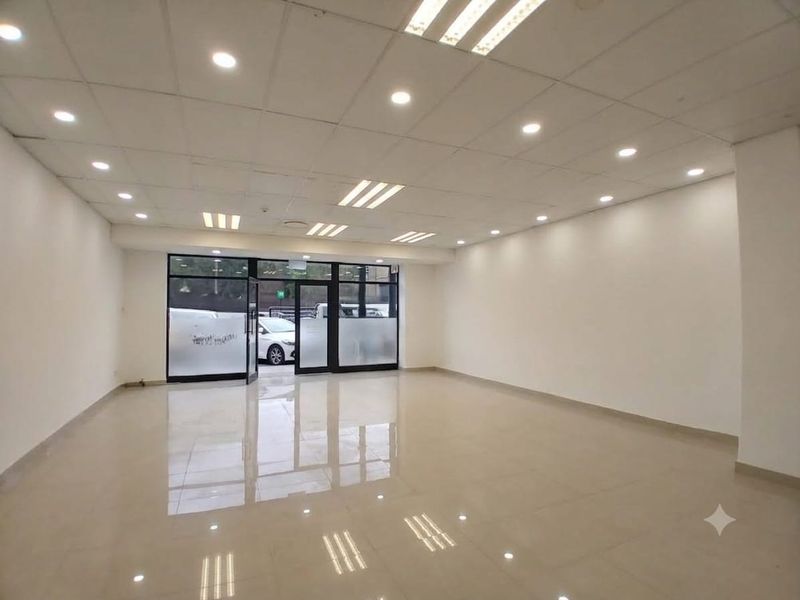A spacious, empty open-plan retail unit featuring polished tiled floors, a suspended ceiling with recessed lighting, and a large glass storefront entrance.