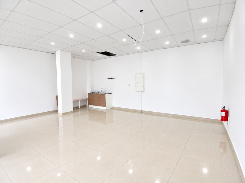 Open plan retail space featuring tiled flooring, suspended ceiling with recessed lighting, a small kitchenette area, and wall-mounted fire safety equipment.