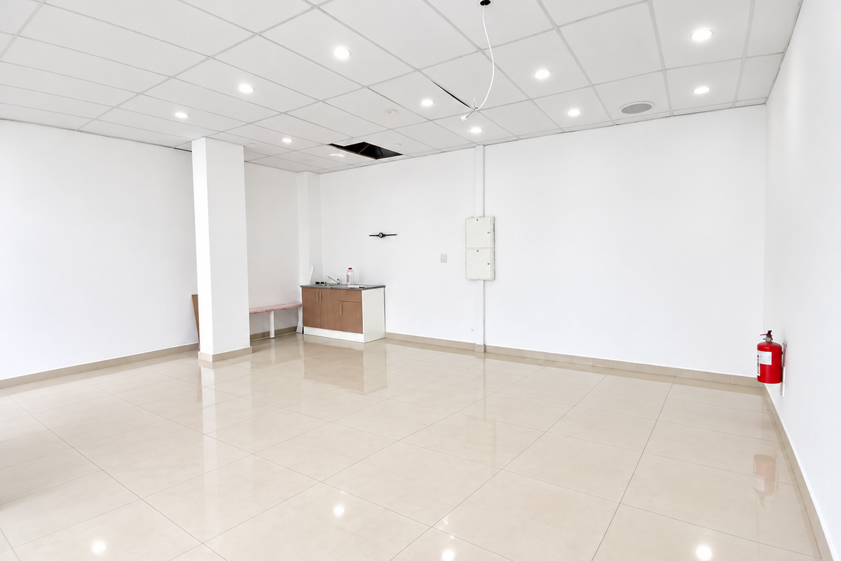 Open plan retail space featuring tiled flooring, suspended ceiling with recessed lighting, a small kitchenette area, and wall-mounted fire safety equipment.