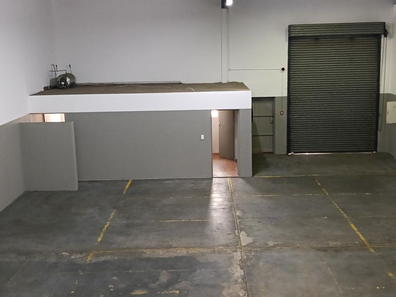 Spacious industrial warehouse unit featuring a high roller shutter door, concrete flooring, and a mezzanine level for additional storage or office space.