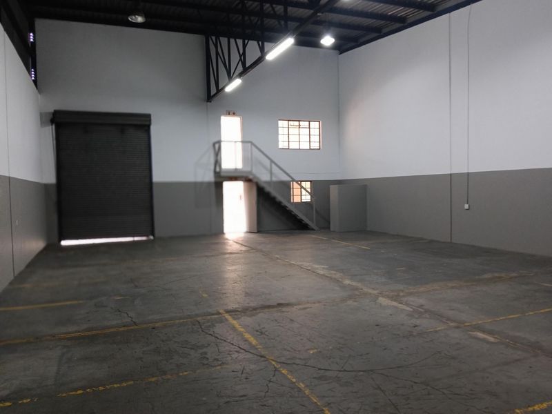 Spacious industrial warehouse unit featuring high ceilings with exposed steel trusses, concrete flooring, a large roller shutter door, and an internal metal staircase.