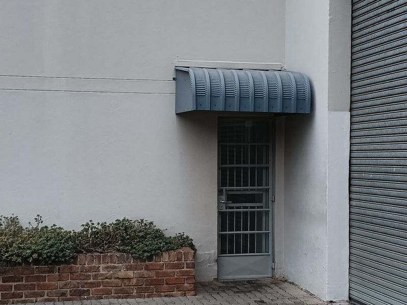 Industrial exterior featuring a roller shutter door, secure pedestrian entrance with metal awning, and a brick planter box on a paved driveway.