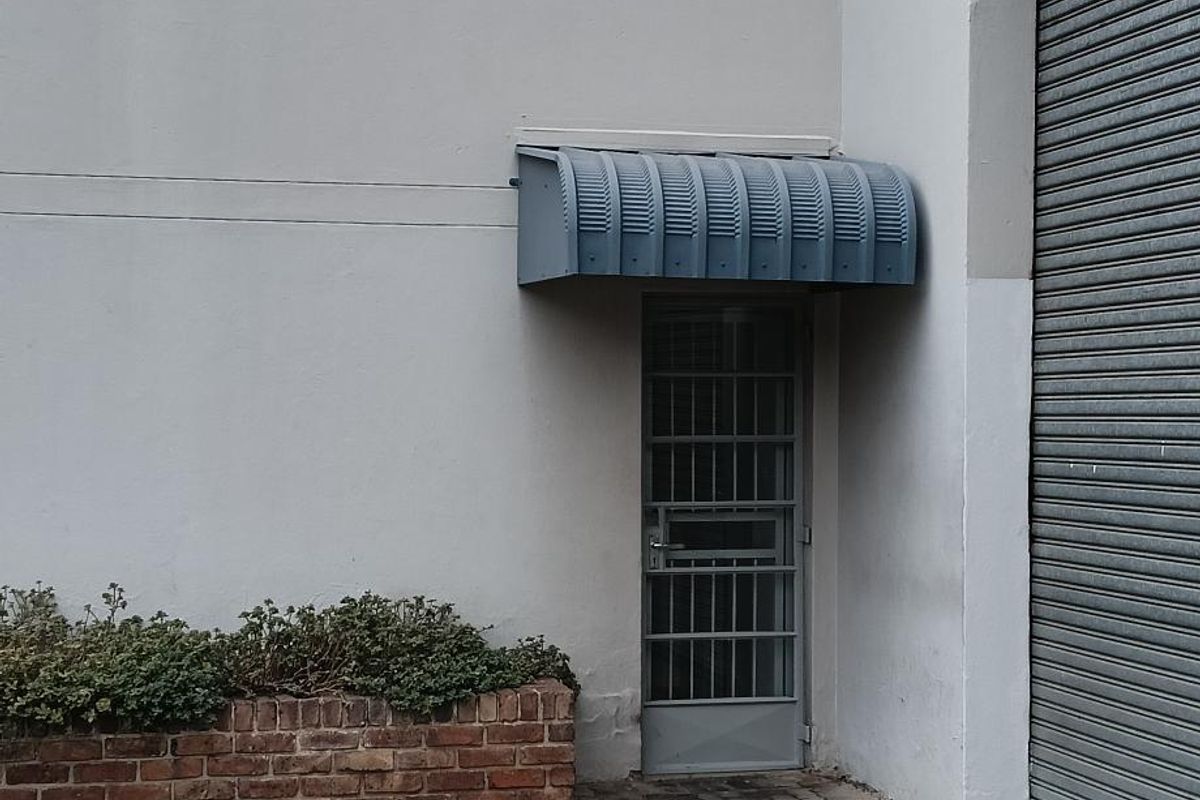 Industrial exterior featuring a roller shutter door, secure pedestrian entrance with metal awning, and a brick planter box on a paved driveway.