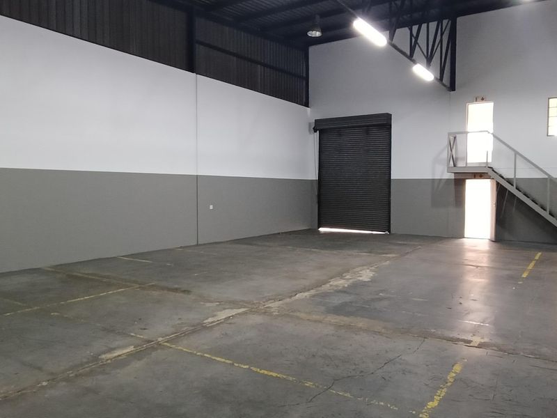 Spacious industrial warehouse unit featuring high ceilings with exposed trusses, a roller shutter door, concrete flooring, and an internal mezzanine staircase.
