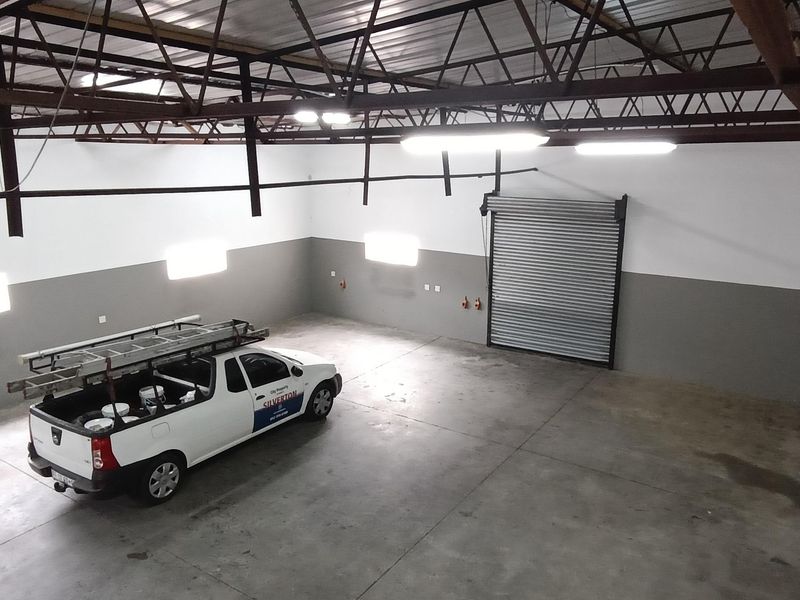 Spacious industrial garage featuring high ceilings with exposed steel trusses, concrete flooring, and a large roller shutter door for easy vehicle access.
