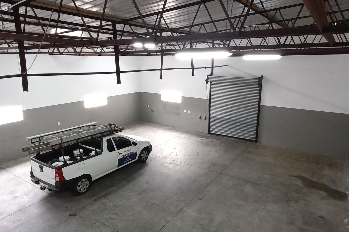 Spacious industrial garage featuring high ceilings with exposed steel trusses, concrete flooring, and a large roller shutter door for easy vehicle access.