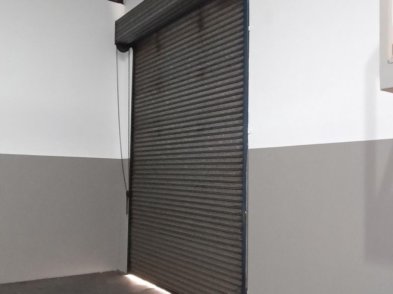 Industrial utility space featuring a secure roller shutter door, concrete flooring, and exposed metal roof structure, suitable for storage or workshop use.