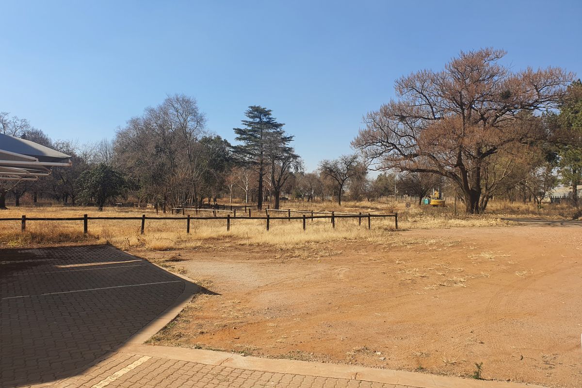 A view of the vacant commercial land featuring a paved parking area, timber fencing, and established trees under a clear sky.