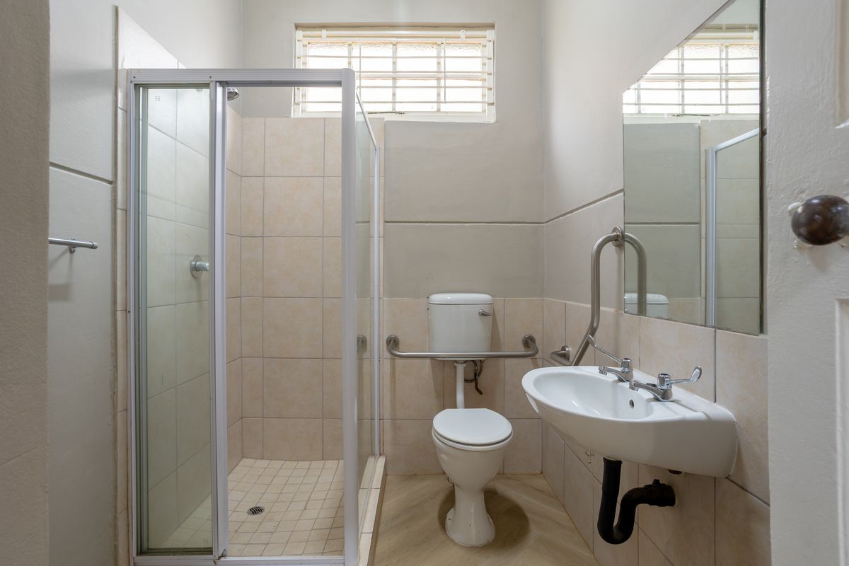 A functional office bathroom featuring a shower enclosure, toilet, wall-mounted basin with grab rails, and a large wall mirror.