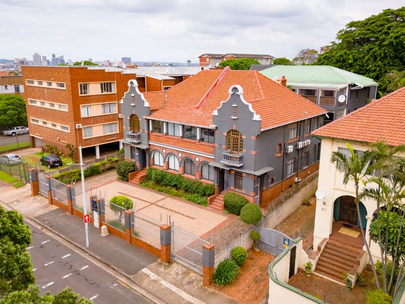 Aerial view of a multi-story office building featuring a distinctive terracotta roof, brick facade, and secure paved parking area in an urban setting.