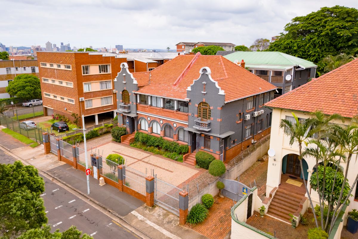 Aerial view of a multi-story office building featuring a distinctive terracotta roof, brick facade, and secure paved parking area in an urban setting.