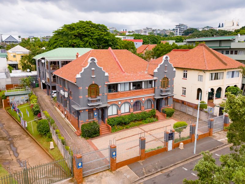 Aerial view of a well-maintained multi-story office building featuring a classic architectural style, tiled roof, paved parking area, and secure perimeter fencing.