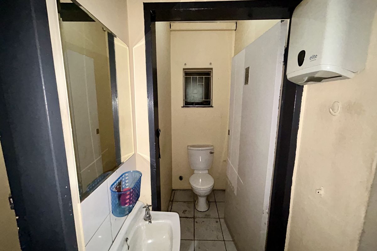 A functional industrial-style bathroom featuring a standard toilet, wall-mounted washbasin, and tiled flooring, requiring cosmetic updates to improve its overall condition.