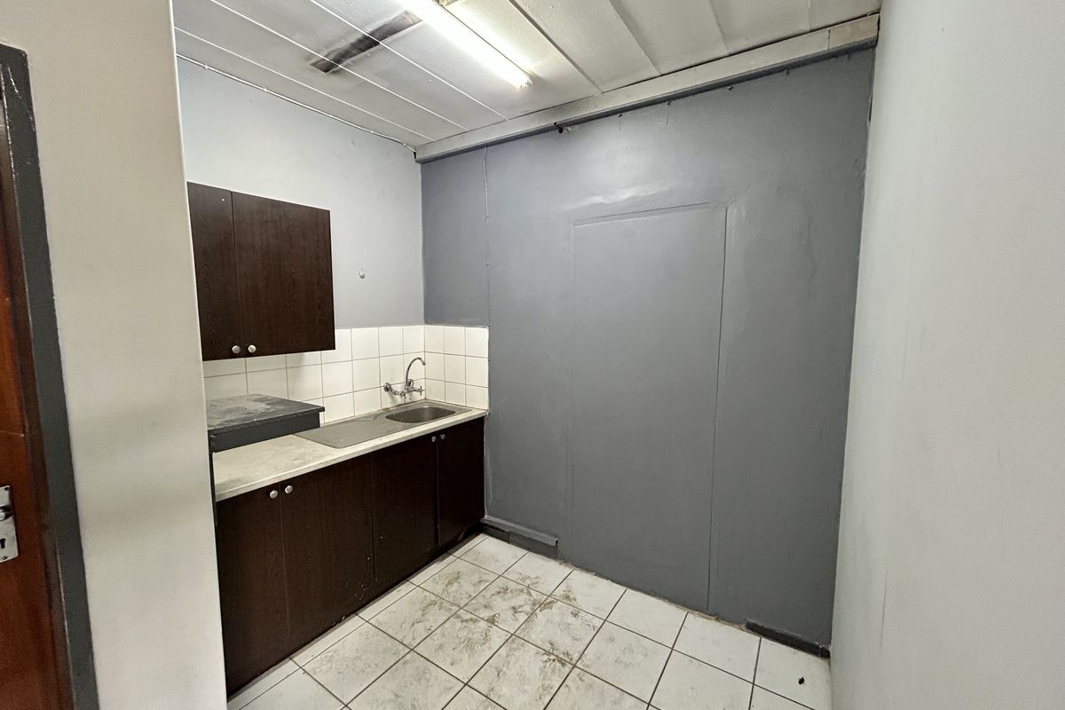 A compact industrial kitchenette featuring dark built-in cabinetry, a stainless steel sink, tiled backsplash, and tiled flooring in need of maintenance.